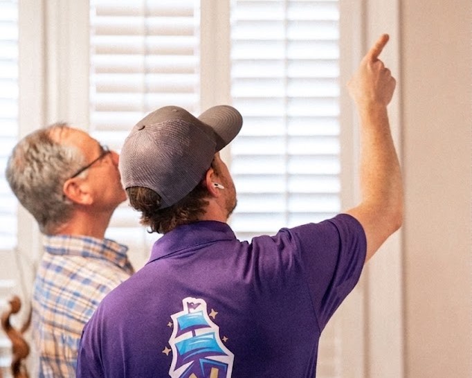 NADCA certified technician pointing up to an air vent while explaining the inspection to a homeowner indoors.